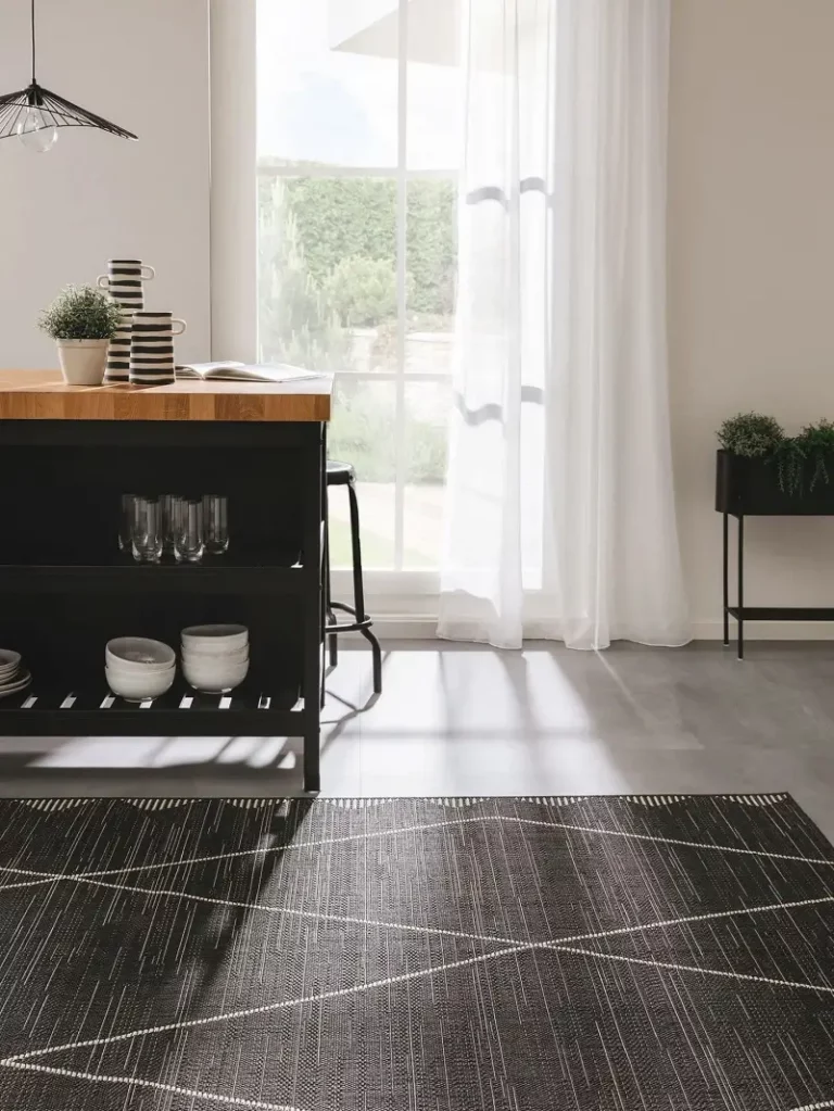 Ein geräumiger Raum mit großen Fenstern, die mit durchsichtigen Vorhängen verhängt sind, einer dunklen Holzanrichte mit Geschirr darunter und einem geometrisch gemusterten dunklen Teppich auf grauem Boden.